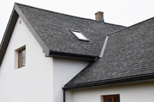 Part of white house with new wooden eaves, plastic rain gutter, window and chimney in grey shingles roof and brown window in facade.