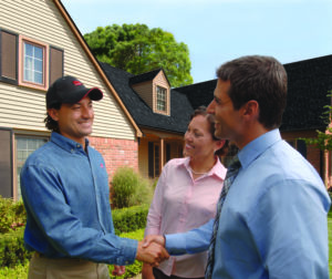 Roofing contractor shaking hands with homeowner. Home with newly installed roof in background.